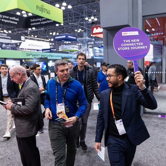 Guided tour at NRF 2025, a speaker holds a sign "The New Connected Store Journey" leading a group through the exhibition area.